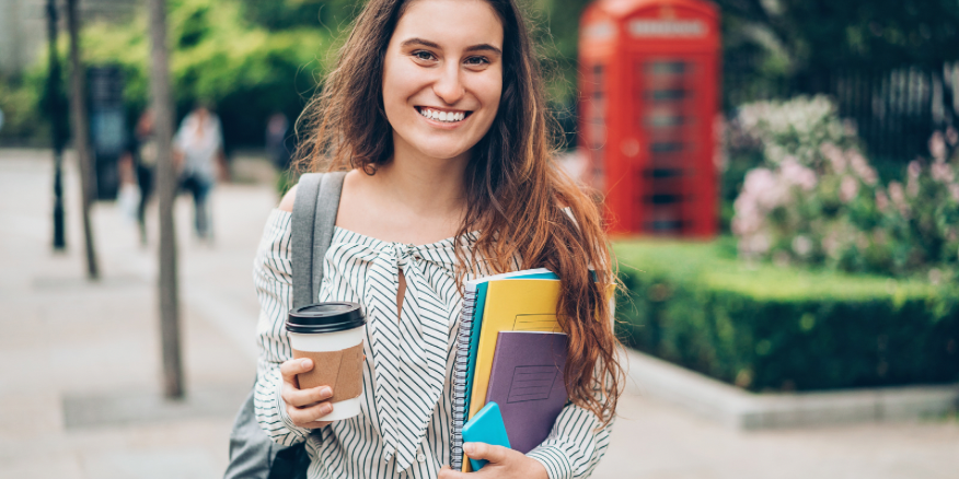 Image of a student expat smiling in the streets of London, holding a coffee and university books